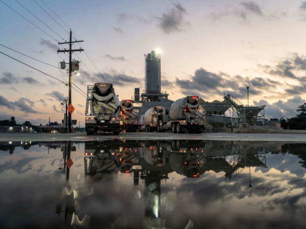 Group of cement mixer trucks at sunset, with puddle reflecting scene.