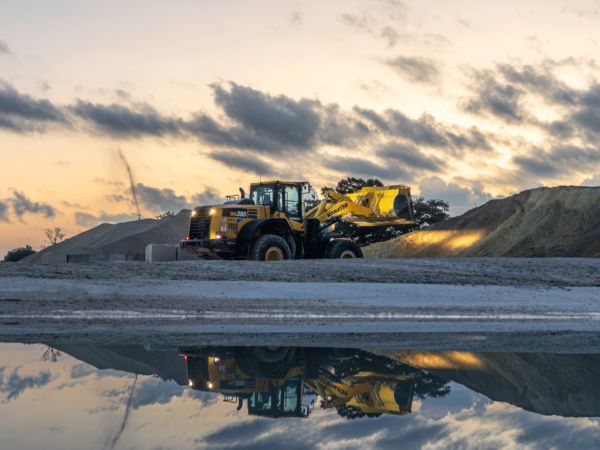 A yellow front loader parked near large mounds of sand or gravel at sunset, with a dramatic sky filled with clouds. The machine and sky are reflected in a puddle on the ground.