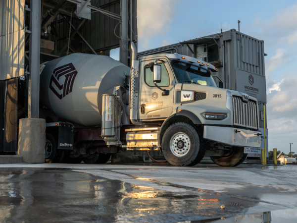 Cement mixer truck getting loaded during the day, with the scene reflected in a puddle.