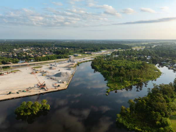 A construction site with various equipment and materials is visible from above, situated along a river bend, with a bridge under construction, and a town and treeline in the distance