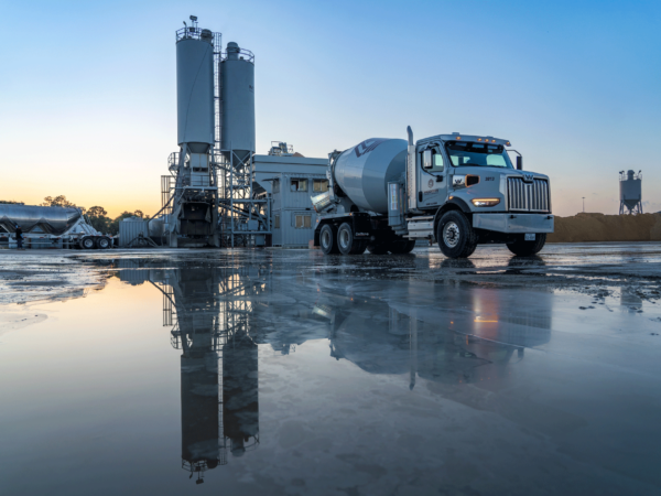 Cement mixer truck at sunrise in front of silos with pool reflecting the scene.