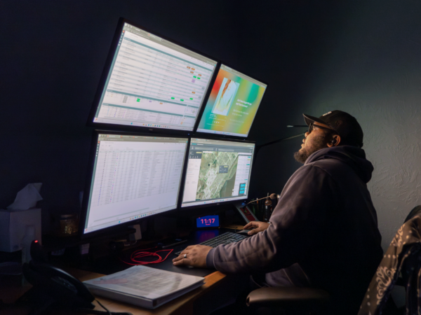 A person wearing a cap and hoodie sitting at a desk with multiple large computer monitors displaying data, maps, and charts in a dark room, illuminated by the glow of the screens.